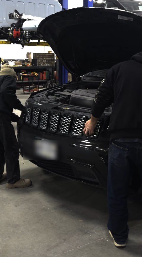 Two technicians working under the open hood of a black Jeep in the service bay