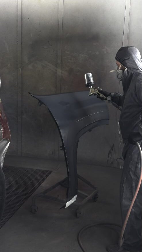 Painter in a hooded suit and respirator spraying a fender inside the paint booth